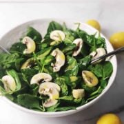 spinach and mushroom salad in a white bowl with a lemon on the side.