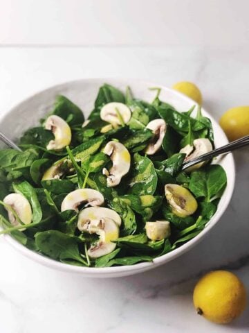 spinach and mushroom salad in a white bowl with a lemon on the side.