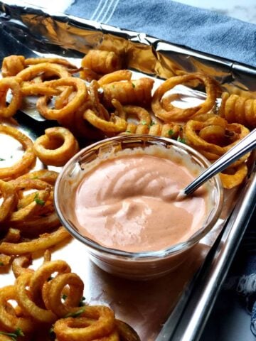 A bowl of sauce sitting on a sheet pan with curly fries.
