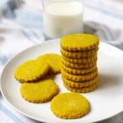 A stack of turmeric cookies on a plate next to a glass of milk.