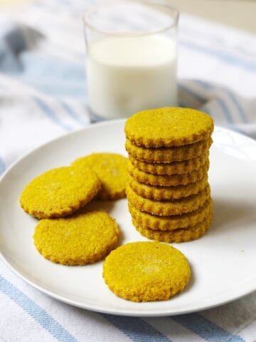 A stack of turmeric cookies on a plate next to a glass of milk.