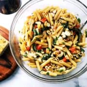 A large bowl of pasta salad on the counter with a glass of wine next to it and bread.