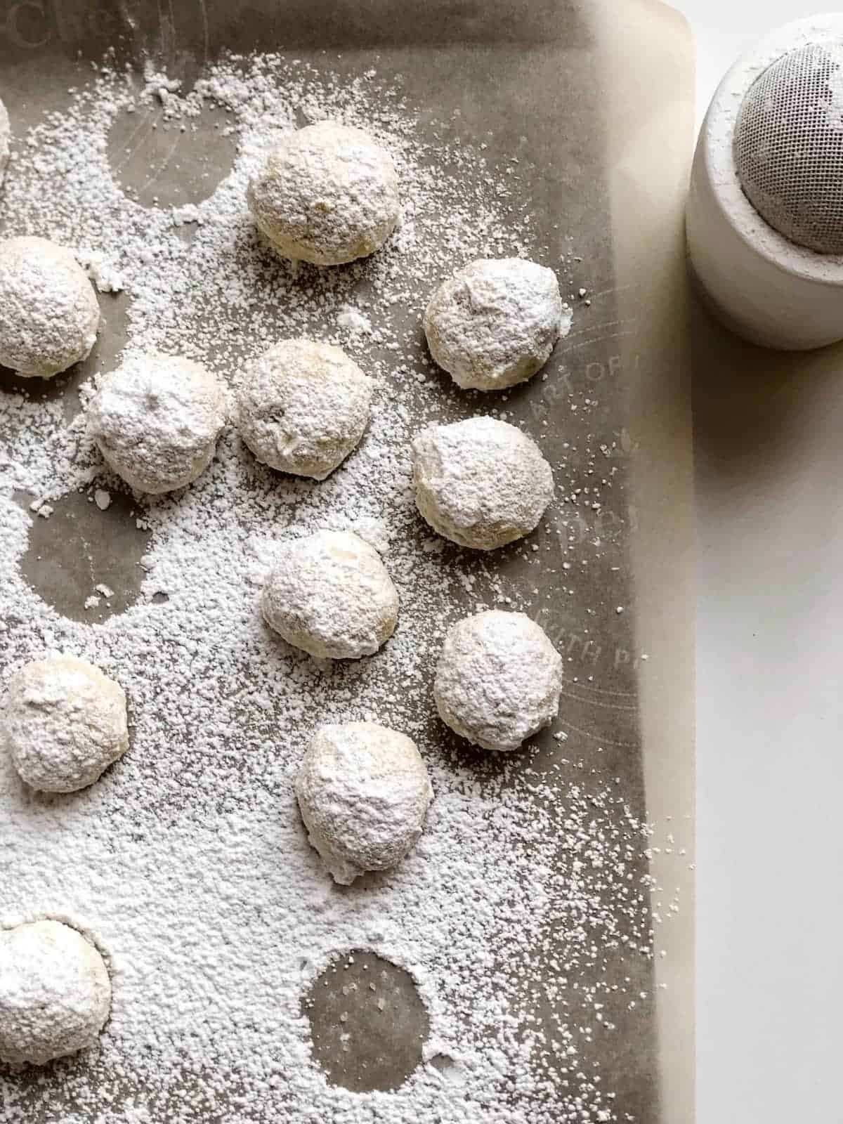 Italian snowball cookies on a sheet pan already baked and powdered sugar being dusted over the top.