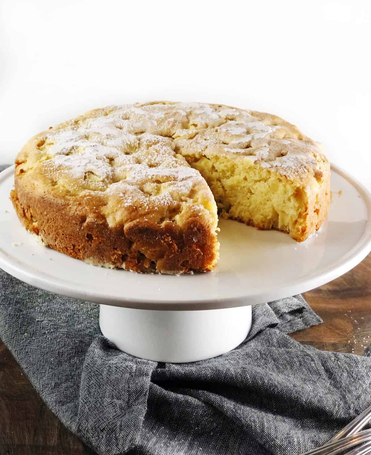 The rustic Italian apple cake on a cake stand with a slice cut out of it.