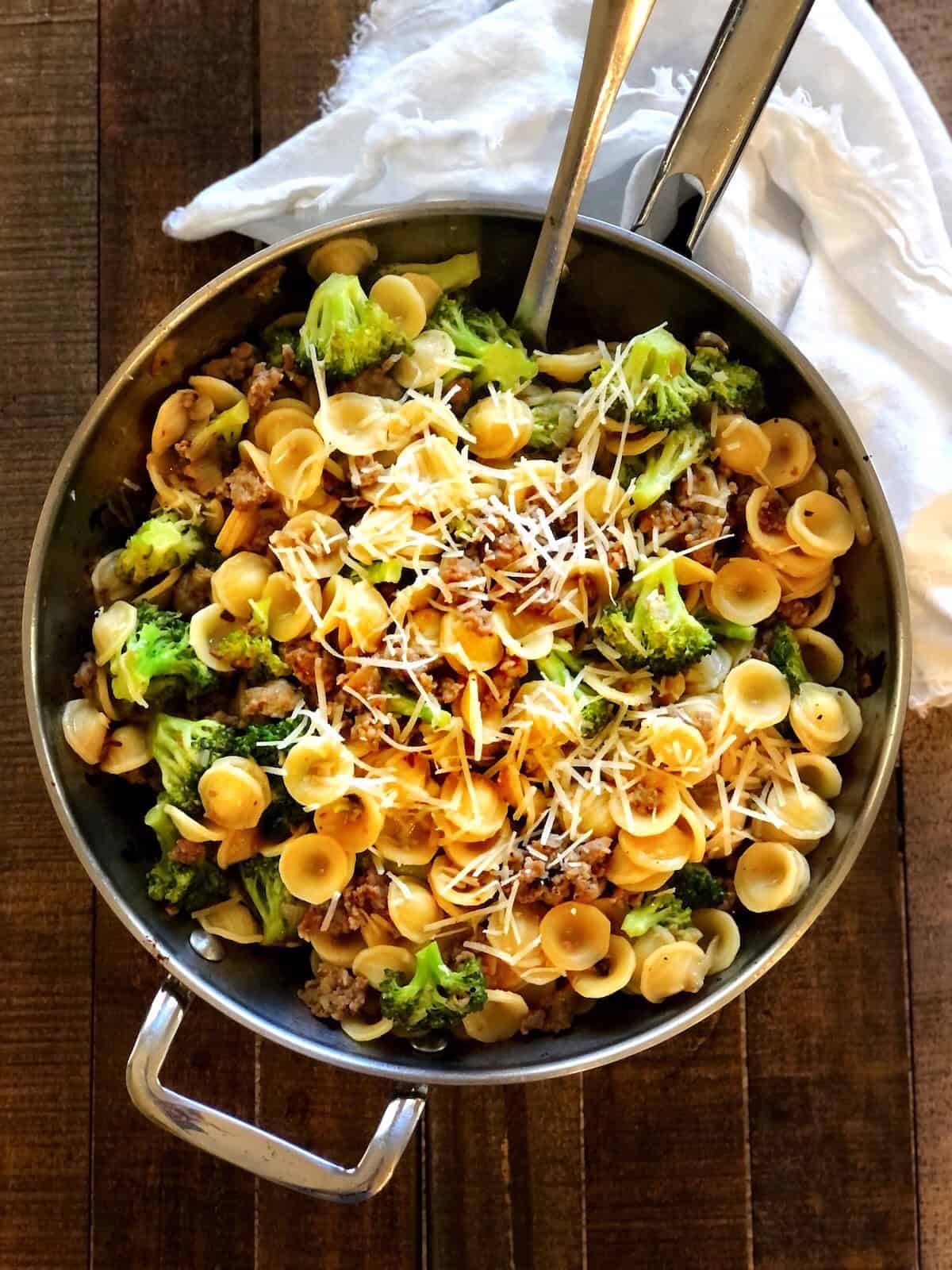 Orecchiette with sausage and broccoli in a large saute pan on a wood table with a cloth napkin next to it.