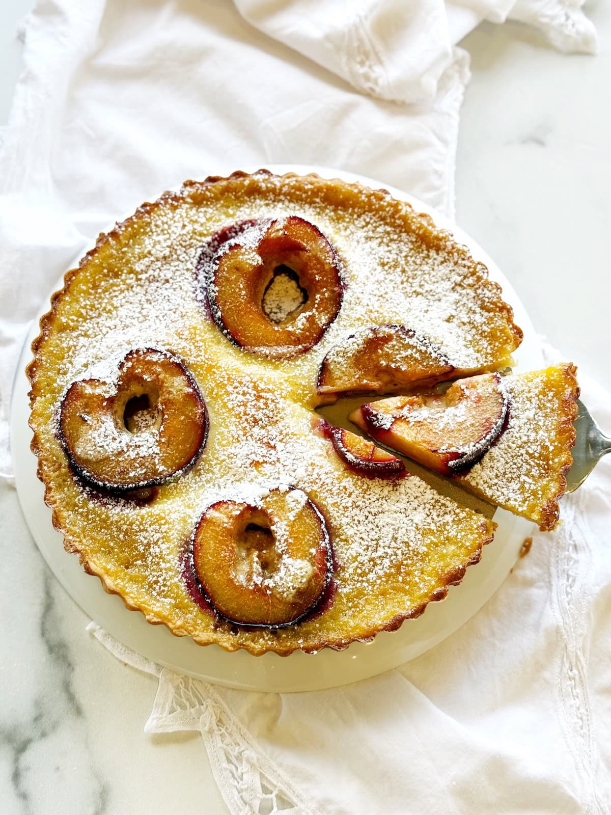 Italian plum and custard tart in a round tart pan on a white counter.